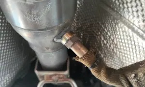 Nahaufnahme einer Lambdasonde, die am Auspuffrohr eines Autos befestigt ist. Sie misst den Sauerstoffgehalt im Abgas, um die optimale Verbrennung zu gewährleisten.