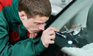 Ein Mechaniker untersucht mit einer Taschenlampe einen Steinschlag an der Windschutzscheibe. Ein Reparaturgerät ist bereits angebracht.