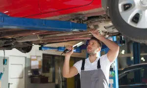 Mechaniker prüft den Unterboden eines roten Autos auf Rost. Er nutzt eine Lampe, um die Stelle für das **unterboden entrosten** besser zu sehen.