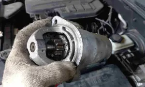 Defekter Anlasser Mercedes: Werkzeug in Handschuh hält ausgebauten Anlasser mit sichtbarem Ritzel. Kosten für Reparatur im Blick.