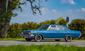 Ein blauer Opel Admiral fährt auf einer Landstraße, umgeben von grünen Bäumen und blauem Himmel.