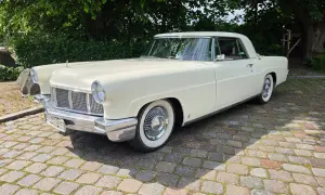 Ein cremefarbener amerikanischer Auto-Klassiker, ein Lincoln Continental, steht auf einem Kopfsteinpflasterweg unter Bäumen.