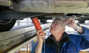 Mechaniker prüft Auto auf Hebebühne mit Lampe. Hoffentlich hat er den Fahrzeugschein dabei, sonst gibt's Ärger!