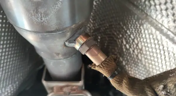 Nahaufnahme einer Lambdasonde, die am Auspuffrohr eines Autos befestigt ist. Sie misst den Sauerstoffgehalt im Abgas, um die optimale Verbrennung zu gewährleisten.