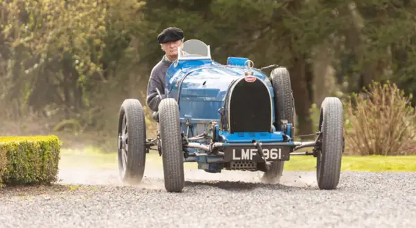 Ein Mann in einem schwarzen Hut fährt einen blauen Bugatti Type 35 über einen Schotterweg.