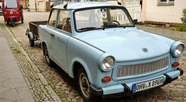 Hellblauer Trabant Kübelwagen mit Anhänger, Baujahr und Zulassung erkennbar.