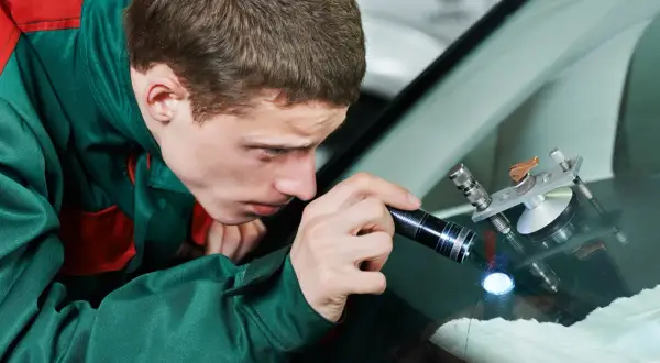 Ein Mechaniker untersucht mit einer Taschenlampe einen Steinschlag an der Windschutzscheibe. Ein Reparaturgerät ist bereits angebracht.