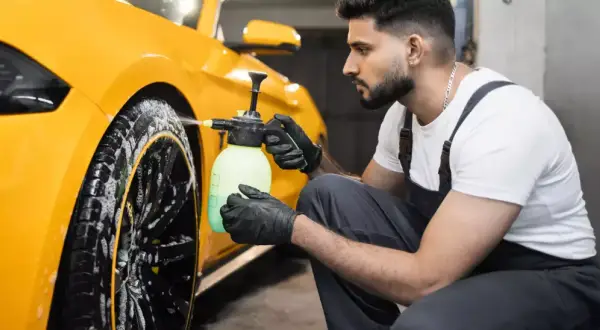 Mann mit Handschuhen sprüht Reinigungsmittel auf gelbe Felgen eines Autos.