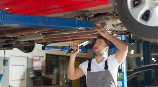 Mechaniker prüft den Unterboden eines roten Autos auf Rost. Er nutzt eine Lampe, um die Stelle für das **unterboden entrosten** besser zu sehen.