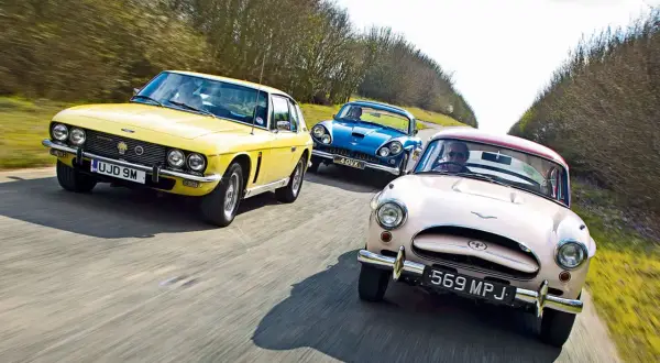 Drei klassische Autos, ein gelber Jensen Interceptor, ein blauer und ein rosafarbener, fahren auf einer Landstraße.