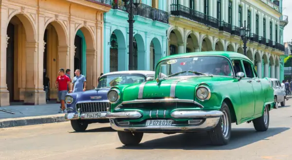 Grüne und blaue Oldtimer fahren auf einer Straße in Havanna. Diese Autos sind ein Symbol für die Geschichte und den Charme der Stadt, die man oft bei einem **oldtimer importieren** erleben kann.