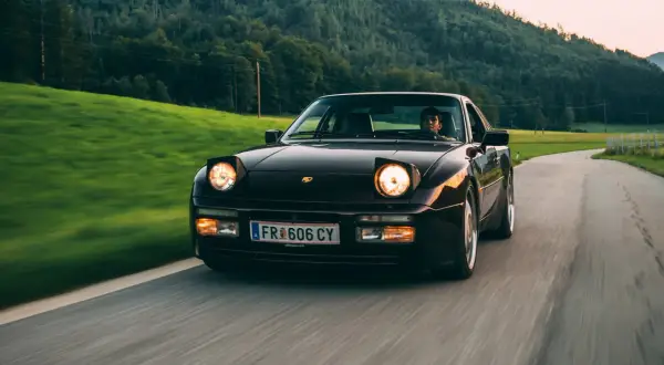 Dunkelroter Porsche mit Klappscheinwerfer fährt auf kurviger Landstraße durch grüne Hügel.