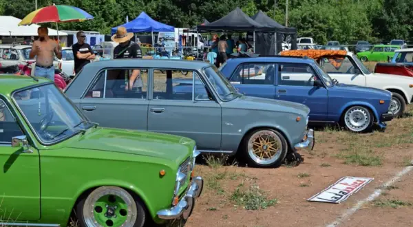 Grüne und graue Lada-Limousinen, ein blauer Lada und weitere Autos stehen auf einem Feld.
