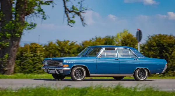 Ein blauer Opel Admiral fährt auf einer Landstraße, umgeben von grünen Bäumen und blauem Himmel.
