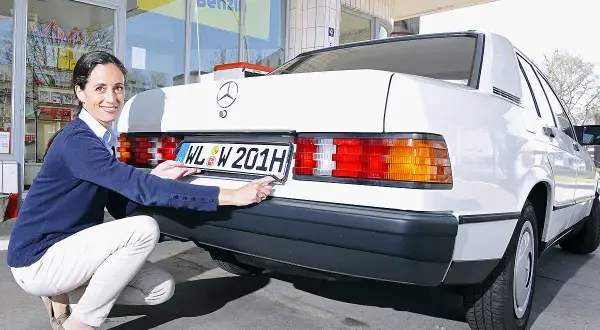 Frau befestigt ein H-Kennzeichen an einem weißen Mercedes. Was kostet ein H Kennzeichen?
