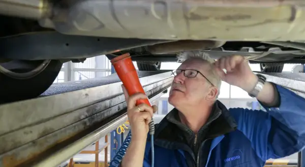 Mechaniker prüft Auto auf Hebebühne mit Lampe. Hoffentlich hat er den Fahrzeugschein dabei, sonst gibt's Ärger!