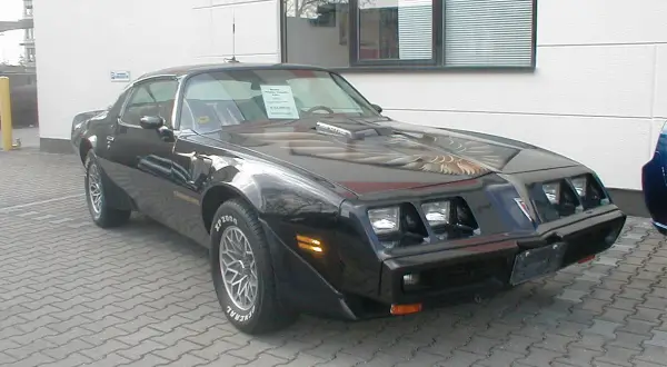Schwarzer Pontiac Trans Am, ein klassisches Muscle Car mit Adler-Grafik auf der Motorhaube, steht auf einem gepflasterten Hof.