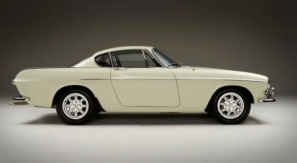 Ein cremefarbener Volvo P1800 Coupé steht im Profil, seine eleganten Linien und Chromdetails glänzen.