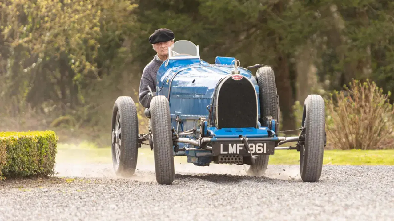 Ein Mann in einem schwarzen Hut fährt einen blauen Bugatti Type 35 über einen Schotterweg.
