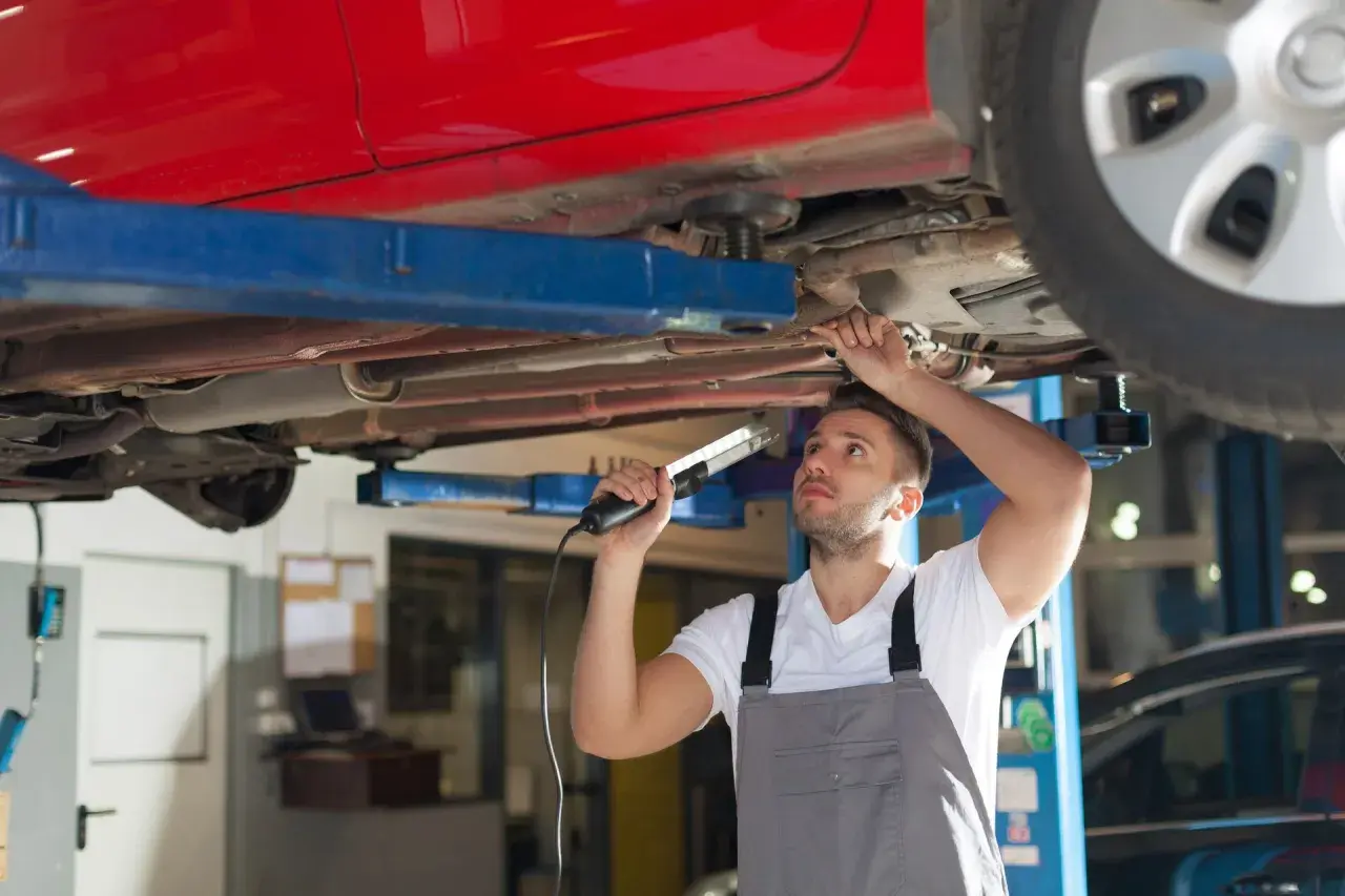 Mechaniker prüft den Unterboden eines roten Autos auf Rost. Er nutzt eine Lampe, um die Stelle für das **unterboden entrosten** besser zu sehen.
