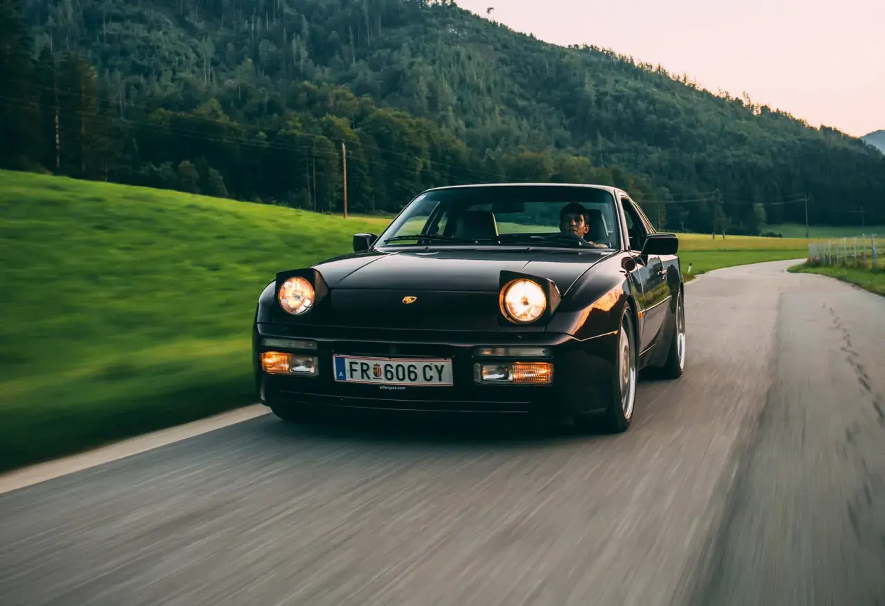 Dunkelroter Porsche mit Klappscheinwerfer fährt auf kurviger Landstraße durch grüne Hügel.