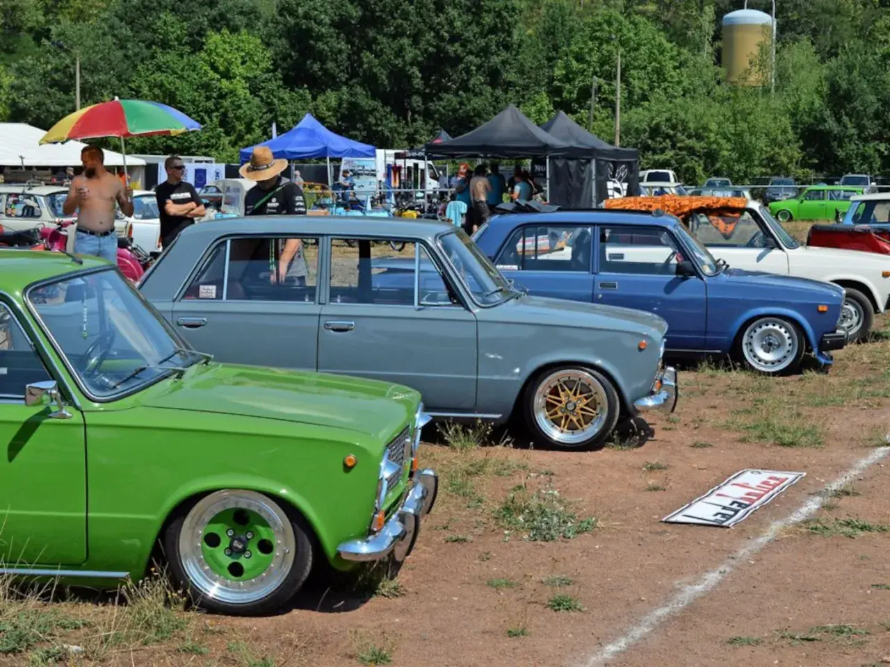 Grüne und graue Lada-Limousinen, ein blauer Lada und weitere Autos stehen auf einem Feld.