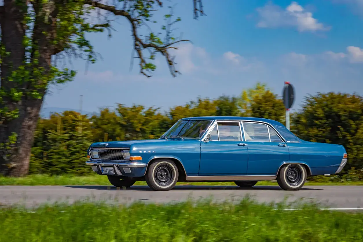 Ein blauer Opel Admiral fährt auf einer Landstraße, umgeben von grünen Bäumen und blauem Himmel.