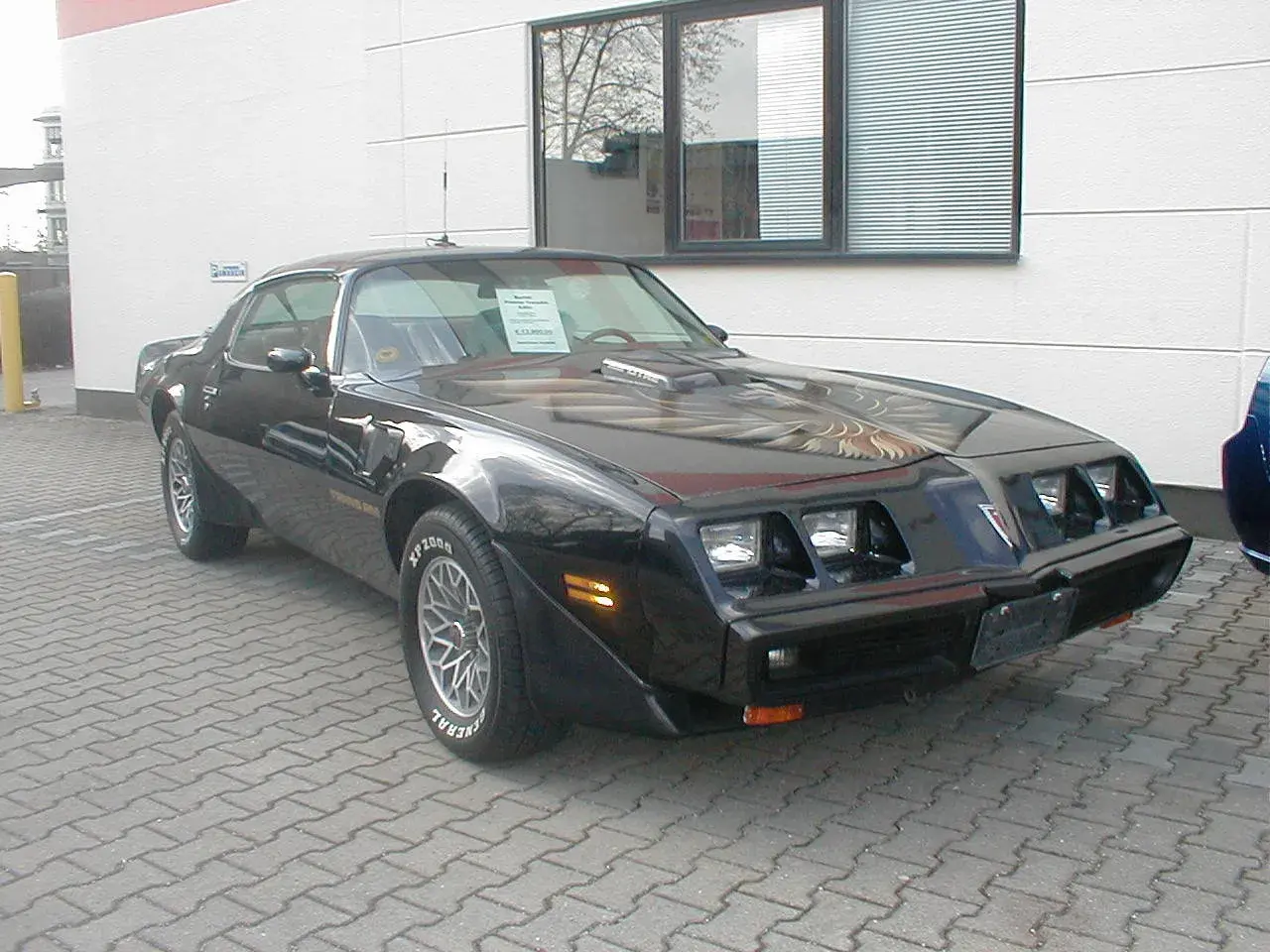 Schwarzer Pontiac Trans Am, ein klassisches Muscle Car mit Adler-Grafik auf der Motorhaube, steht auf einem gepflasterten Hof.