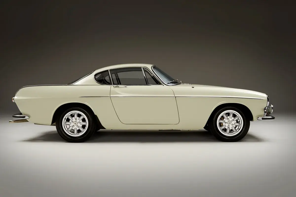 Ein cremefarbener Volvo P1800 Coupé steht im Profil, seine eleganten Linien und Chromdetails glänzen.
