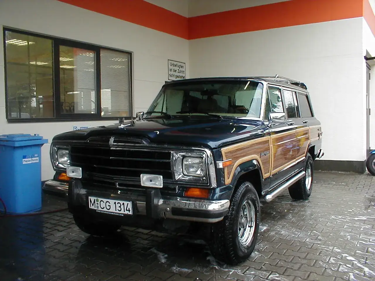 Ein klassisches amerikanisches Auto, ein dunkelblauer Jeep Grand Wagoneer mit Holzverkleidung, steht auf einem nassen, gepflasterten Untergrund.
