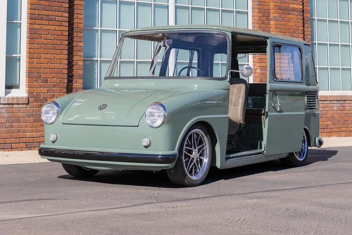 Ein gr&uuml;ner VW Fridolin mit ge&ouml;ffneter T&uuml;r steht vor einer Backsteinwand mit gro&szlig;en Fenstern.