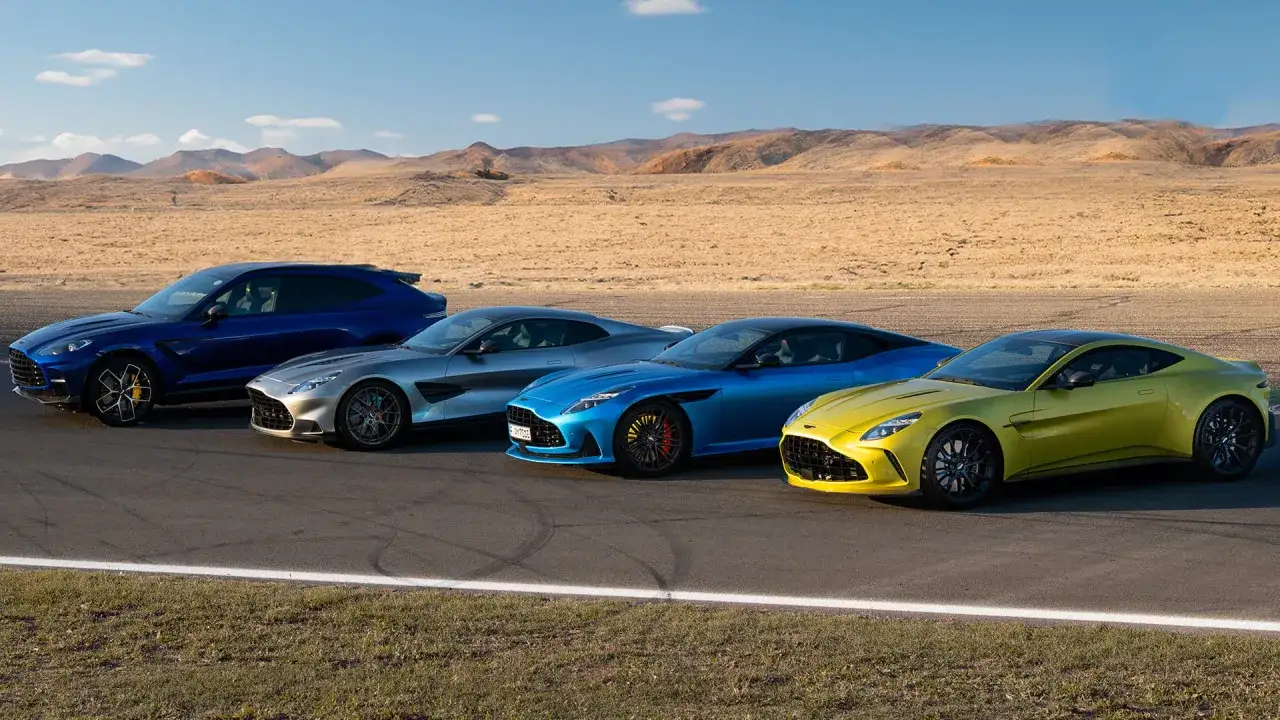 Vier Aston Martin Sportwagen in verschiedenen Farben stehen nebeneinander auf einer Rennstrecke vor einer Wüstenlandschaft.