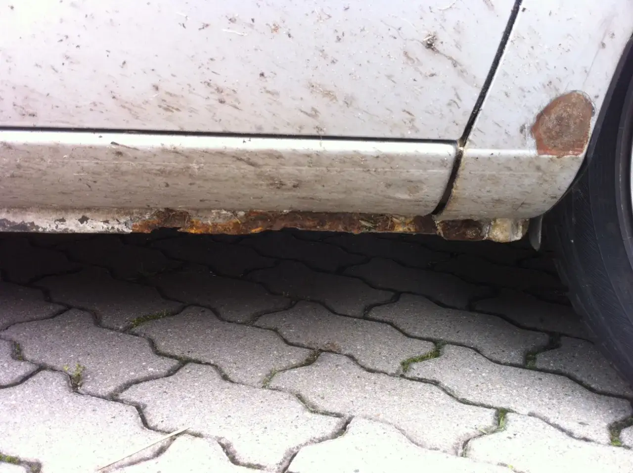 Detailaufnahme zeigt, wie entsteht rost am auto: stark verrostete Schweller und Radlauf an einem silbernen Fahrzeug.
