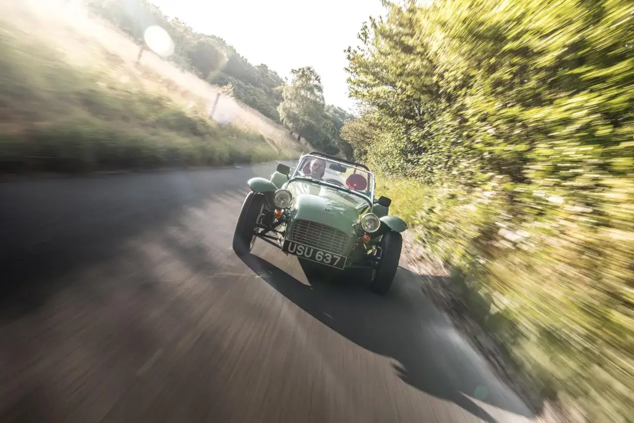 Gr&uuml;ner englischer Roadster auf kurviger Landstra&szlig;e, ein Fahrer im offenen Cockpit, die Landschaft verschwimmt im Fahrtwind.