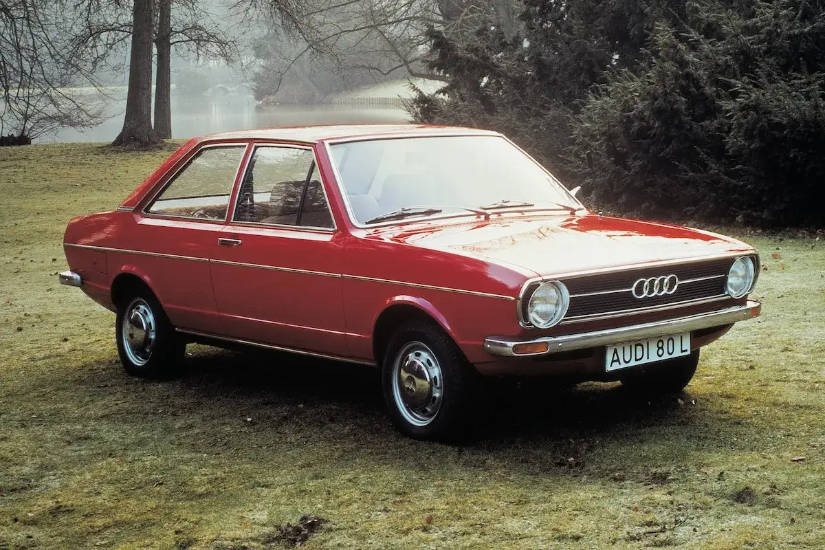 Roter Audi 80 L von 1970 parkt auf einer Wiese, im Hintergrund Bäume und ein Teich.