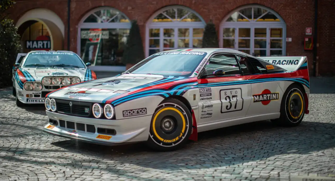 Zwei Lancia Rally 037 mit Martini Racing Lackierung stehen nebeneinander. Der vordere, eine Kimera Evo 37, ist detailreich mit Sponsorenlogos und der Startnummer 37 versehen.