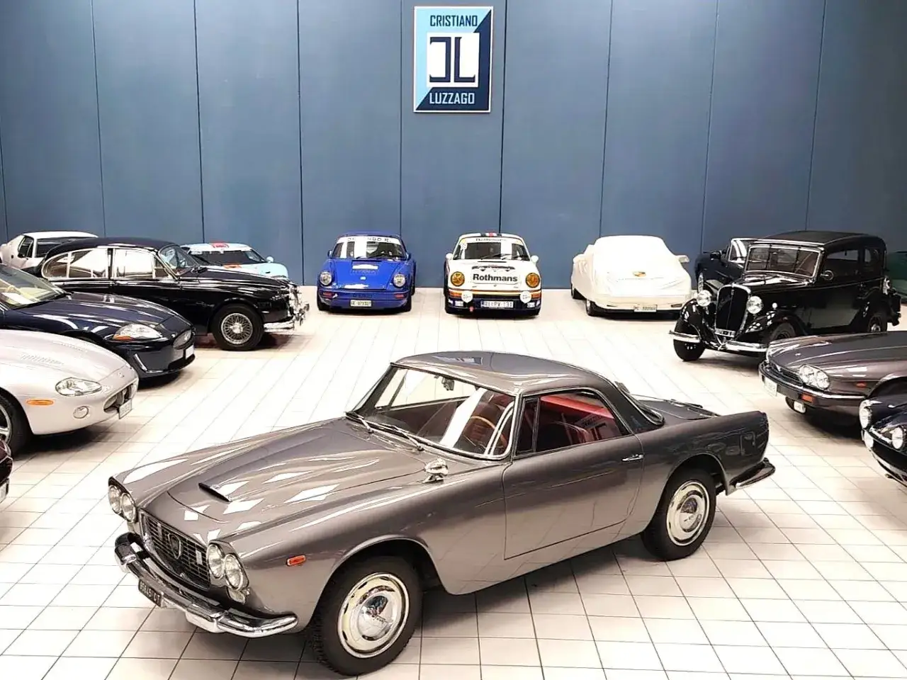 Grauer Lancia Flaminia Coup&eacute; mit rotem Interieur, umgeben von anderen Oldtimern in einer Ausstellungshalle.