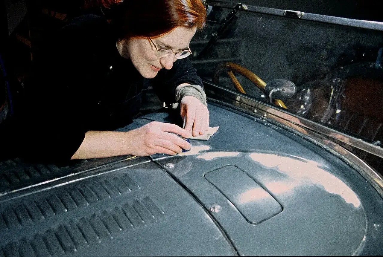 Frau mit Brille schleift sorgf&auml;ltig an der Motorhaube eines Oldtimers, um die Patina-Lackierung zu erhalten.