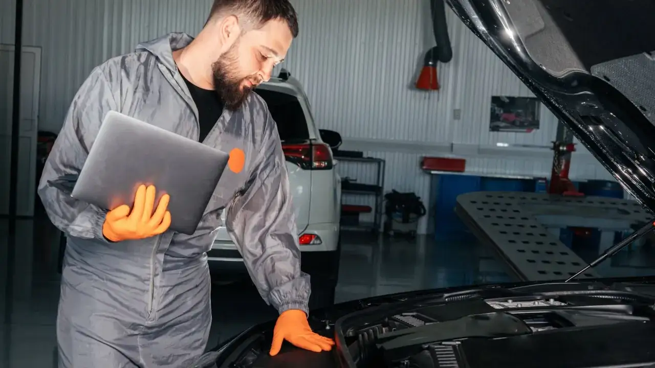 Mechaniker mit Laptop pr&uuml;ft den Motorraum. Er informiert sich &uuml;ber die keilriemen wechseln kosten, bevor er mit der Reparatur beginnt.