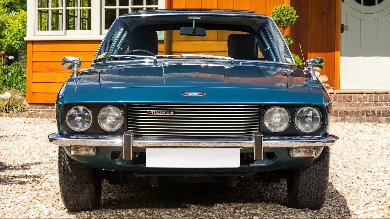 Ein klassisches Jensen Auto, ein Interceptor II in tiefem Blau, steht vor einem charmanten Haus.