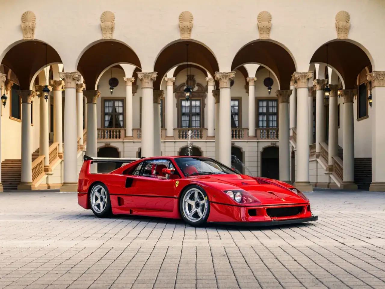 Roter Ferrari F40 LM vor einer pr&auml;chtigen Villa mit S&auml;uleng&auml;ngen. Ein Traumauto auf gepflastertem Hof.