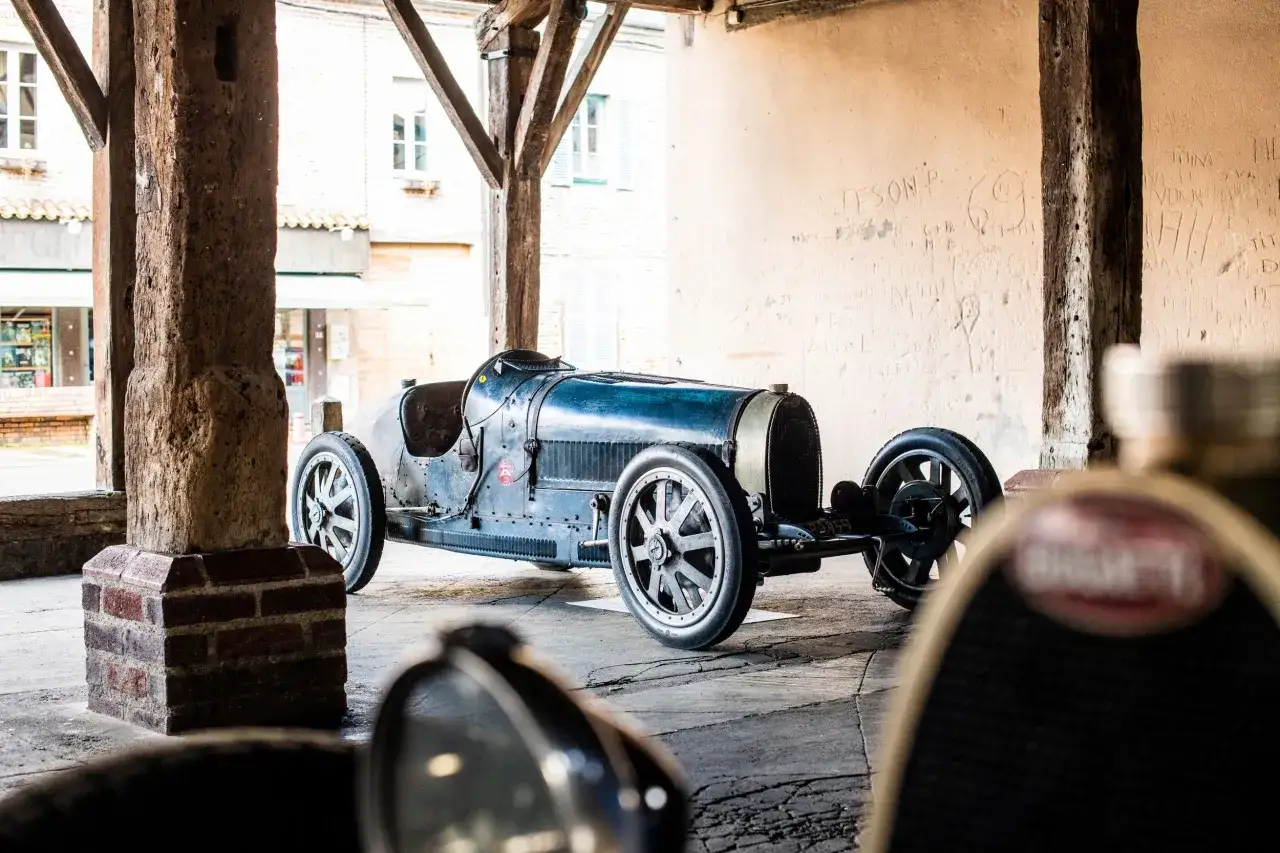 Ein alter Bugatti, ein Meisterwerk der Ingenieurskunst, steht unter einem Holzdach. Seine blaue Karosserie gl&auml;nzt im Licht.
