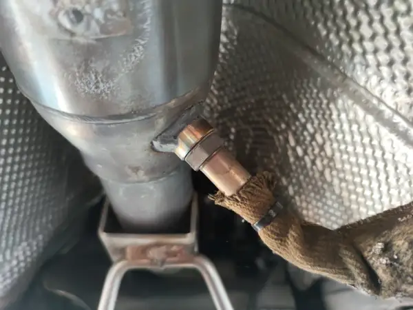 Nahaufnahme einer Lambdasonde, die am Auspuffrohr eines Autos befestigt ist. Sie misst den Sauerstoffgehalt im Abgas, um die optimale Verbrennung zu gewährleisten.