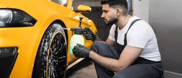 Mann mit Handschuhen sprüht Reinigungsmittel auf gelbe Felgen eines Autos.