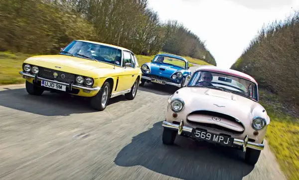 Drei klassische Autos, ein gelber Jensen Interceptor, ein blauer und ein rosafarbener, fahren auf einer Landstraße.