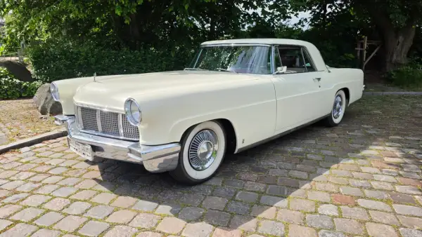 Ein cremefarbener amerikanischer Auto-Klassiker, ein Lincoln Continental, steht auf einem Kopfsteinpflasterweg unter Bäumen.