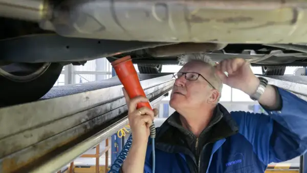 Mechaniker prüft Auto auf Hebebühne mit Lampe. Hoffentlich hat er den Fahrzeugschein dabei, sonst gibt's Ärger!